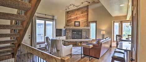 A spacious living area with a fireplace, wooden flooring, a high ceiling with a fan, and a staircase with a railing.