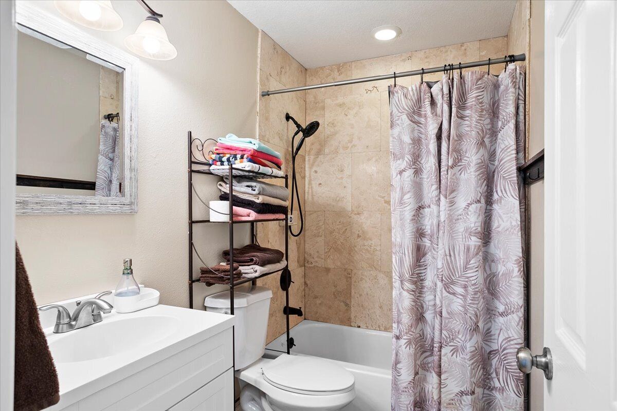 Une salle de bain avec un lavabo, des toilettes et une baignoire, tous de couleur blanche. On y trouve un porte-serviettes avec des serviettes pliées et une pomme de douche.