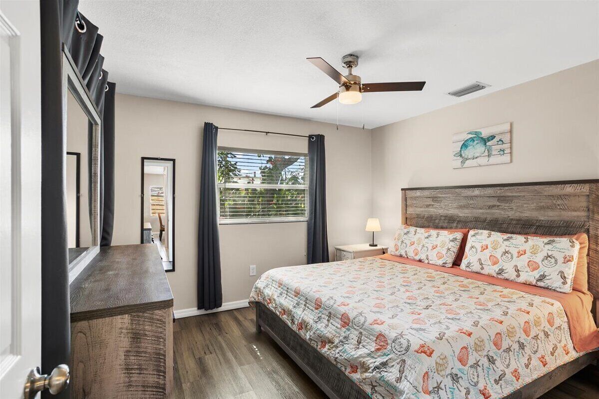 Ein Schlafzimmer mit einem Bett, einem Deckenventilator, einem Fenster mit Vorhängen, einem Nachttisch mit Lampe und einem gerahmten Bild an der Wand.
