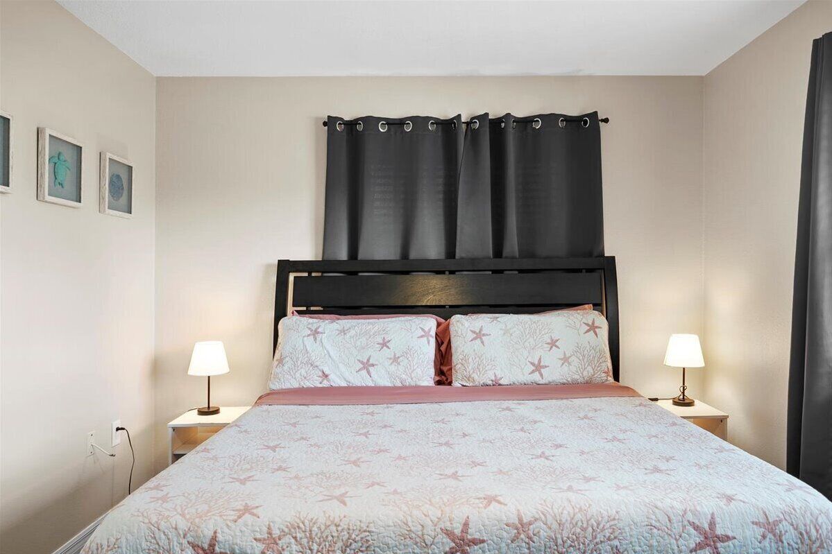 A neatly made bed with a floral bedspread, two bedside lamps, and a dark curtain over the window.