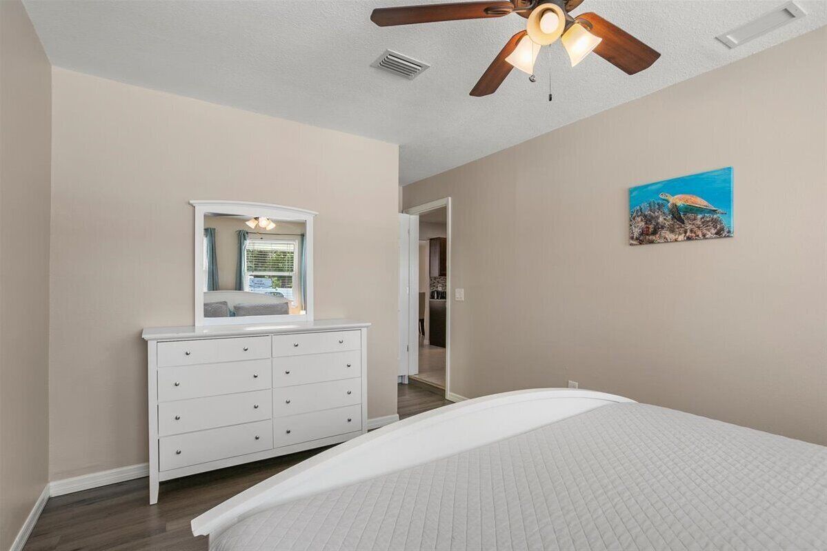 A bedroom with a bed, a dresser, a ceiling fan, and a painting on the wall.