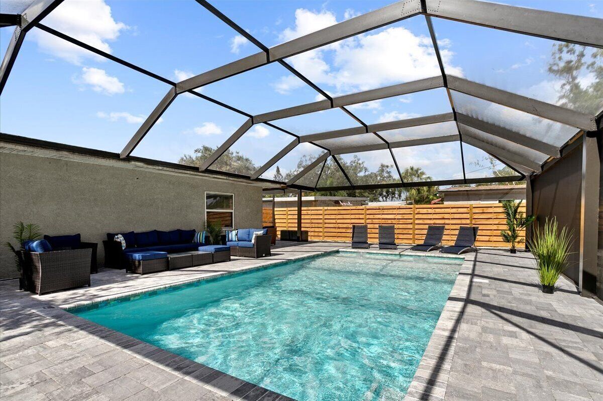 Une piscine extérieure couverte, dotée de chaises longues et de canapés, entourée d’une clôture en bois et agrémentée de plantes en pot.