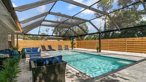 An outdoor swimming pool with a covered patio area, lounge chairs, and a wooden fence.