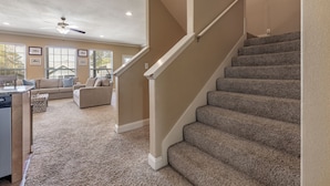 Un intérieur de maison comprenant un escalier recouvert de moquette, un ventilateur de plafond et un espace de vie avec un canapé et une table à manger.