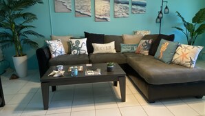 A living room with a sectional sofa, a coffee table, and a wall decorated with beach-themed artwork. - Tropical one bedroom condo steps from pool. (Christiansted)