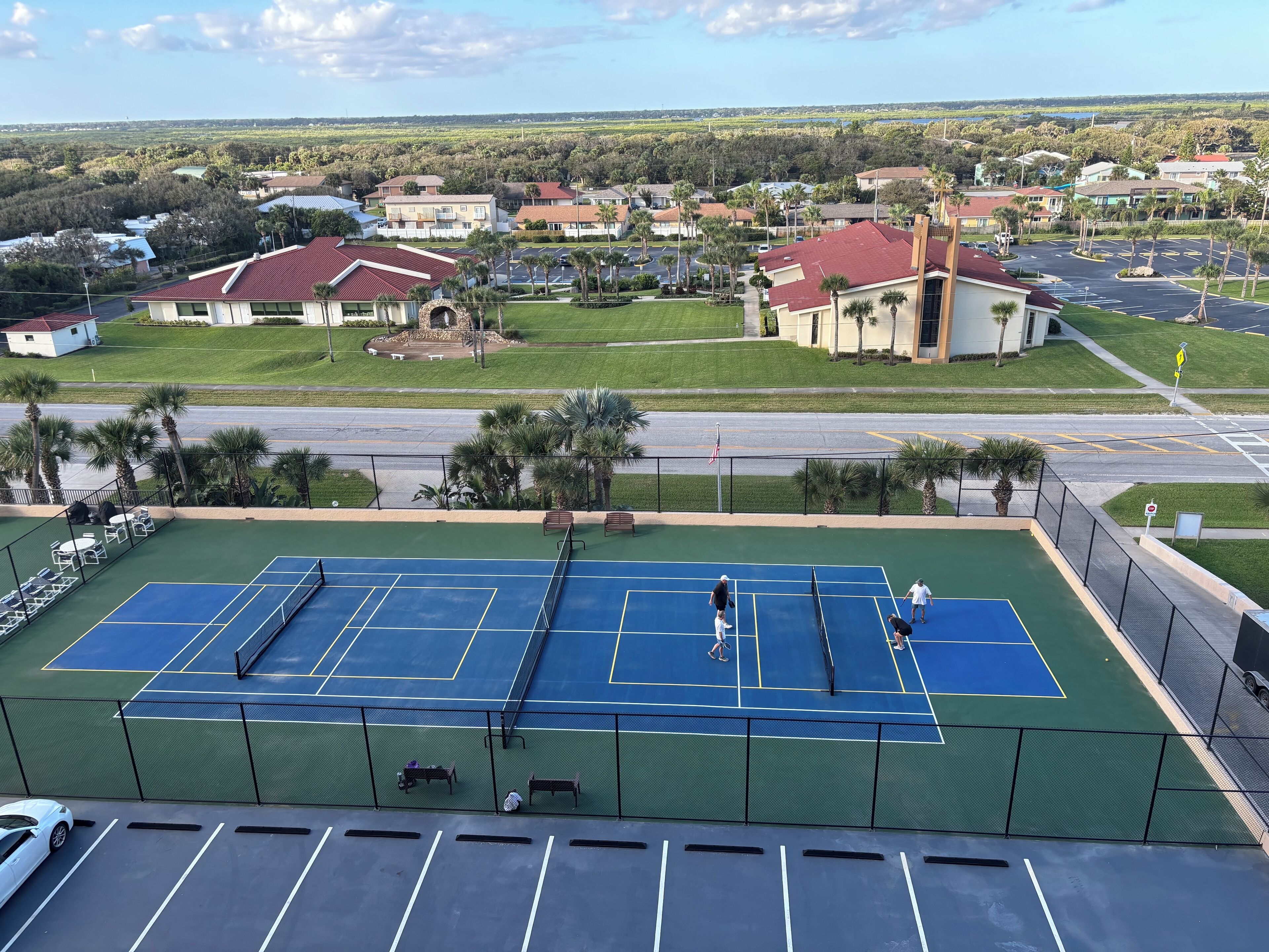 Een tennisbaan met een groen en blauw oppervlak, omringd door een hek, met een parkeerplaats op de voorgrond en huizen op de achtergrond.
