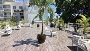 A rooftop terrace with outdoor seating, palm trees, and a modern building in the background. - Amazing view at Riviera Colonial & zona Colonial (Villa Duarte)