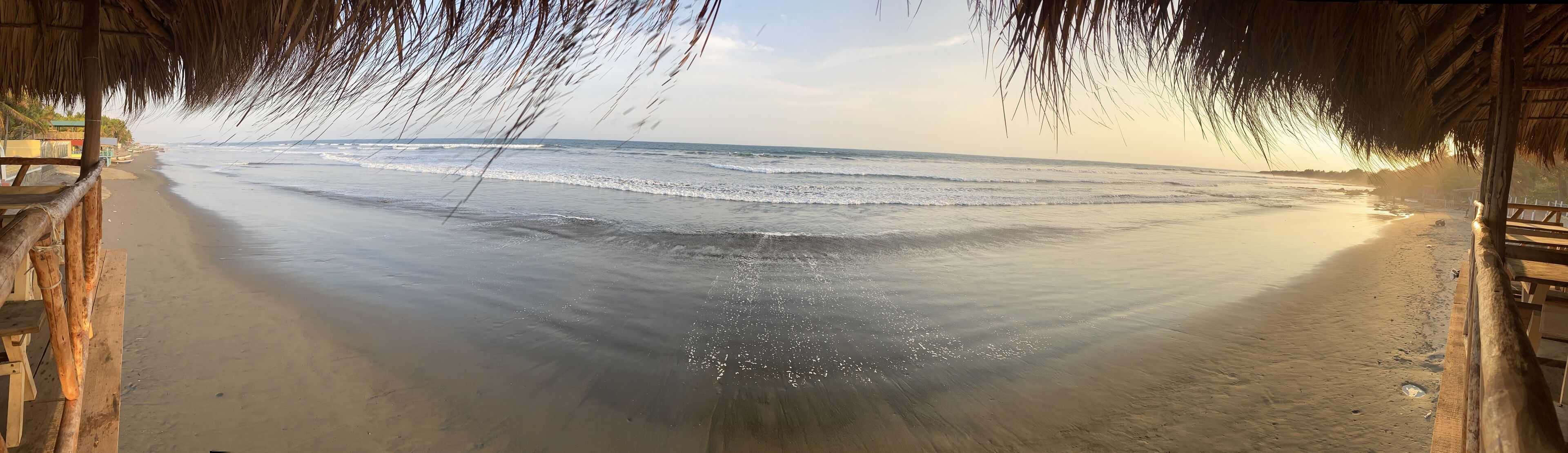 Una playa con un paseo de madera que lleva a la orilla, rodeada de palmeras y una estructura con techo de paja.