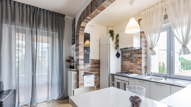 Een moderne keuken met een bakstenen muur, witte kasten, een groot raam en een eethoek met een tafel en stoelen.