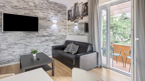 Een moderne woonkamer met een wand met stenen patroon, een flatscreen televisie, een leren bank, een klein tafeltje met een plant, en een balkon met stoelen.