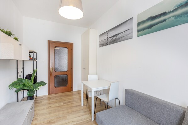 A room with a wooden floor, white walls, a white table with chairs, a grey sofa, a door with a frosted glass panel, and a plant.