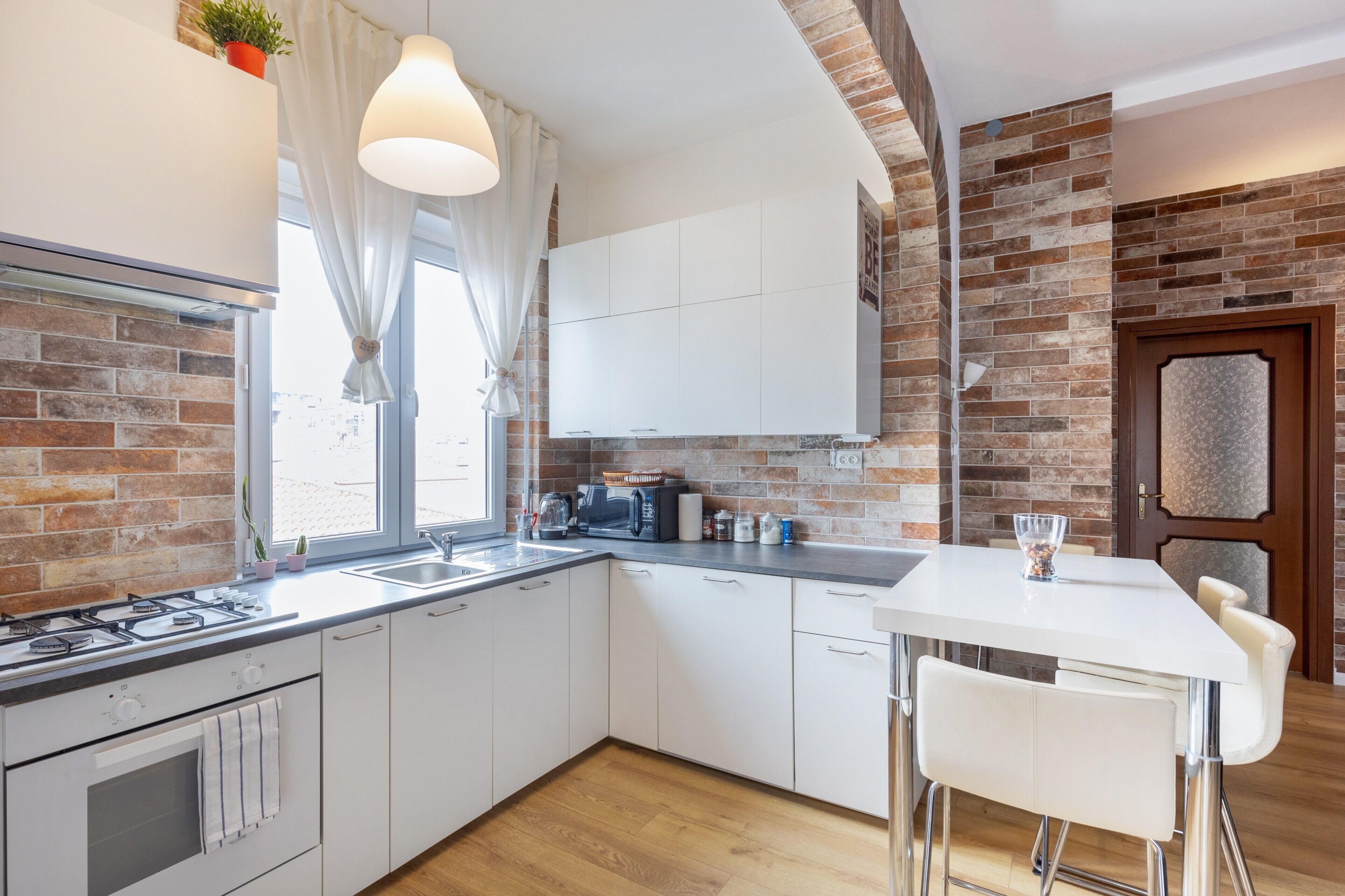Una cucina moderna con mobili bianchi, una parete decorata in mattoni, un'area pranzo con tavolo e sedie bianche e una finestra con tende.