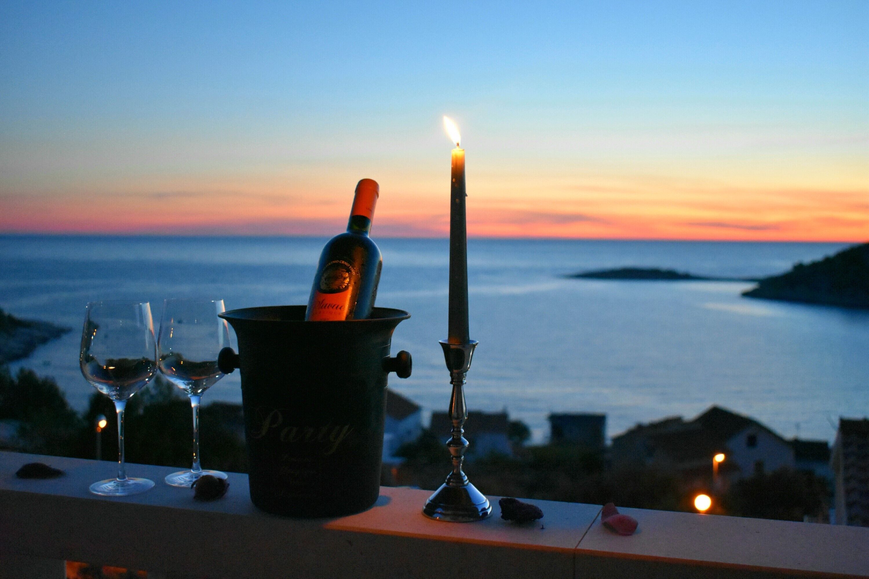 A candle, two wine glasses, and a bottle of wine on a balcony overlooking a body of water at sunset.