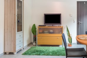 Une pièce avec un téléviseur posé sur un meuble en bois, un tapis vert, une table à manger avec des chaises et une porte.