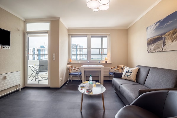 A living room with a sofa, a coffee table with a bottle and glasses, a dining table with chairs, and a large window with a city view.