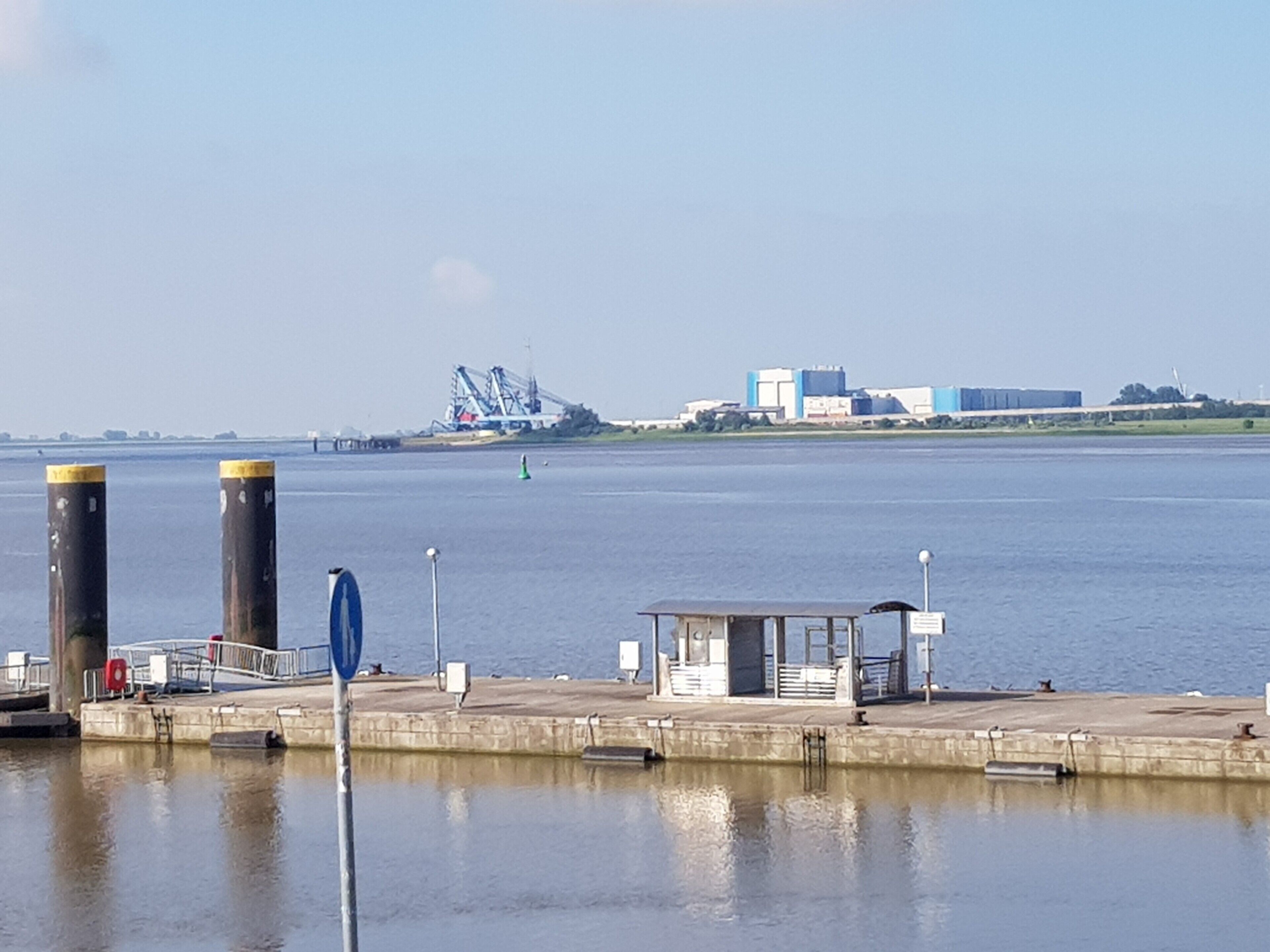 Blick auf das Wasser mit einem Steg, einer kleinen Schutzhütte und Industriegebäuden im Hintergrund.
