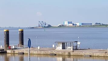 Blick auf das Wasser mit einem Steg, einer kleinen Schutzhütte und Industriegebäuden im Hintergrund.