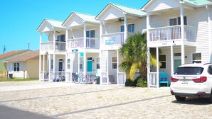 Une rangée d’hôtels blancs à deux étages, situés en bord de mer et dotés de balcons, avec une voiture garée et un palmier.