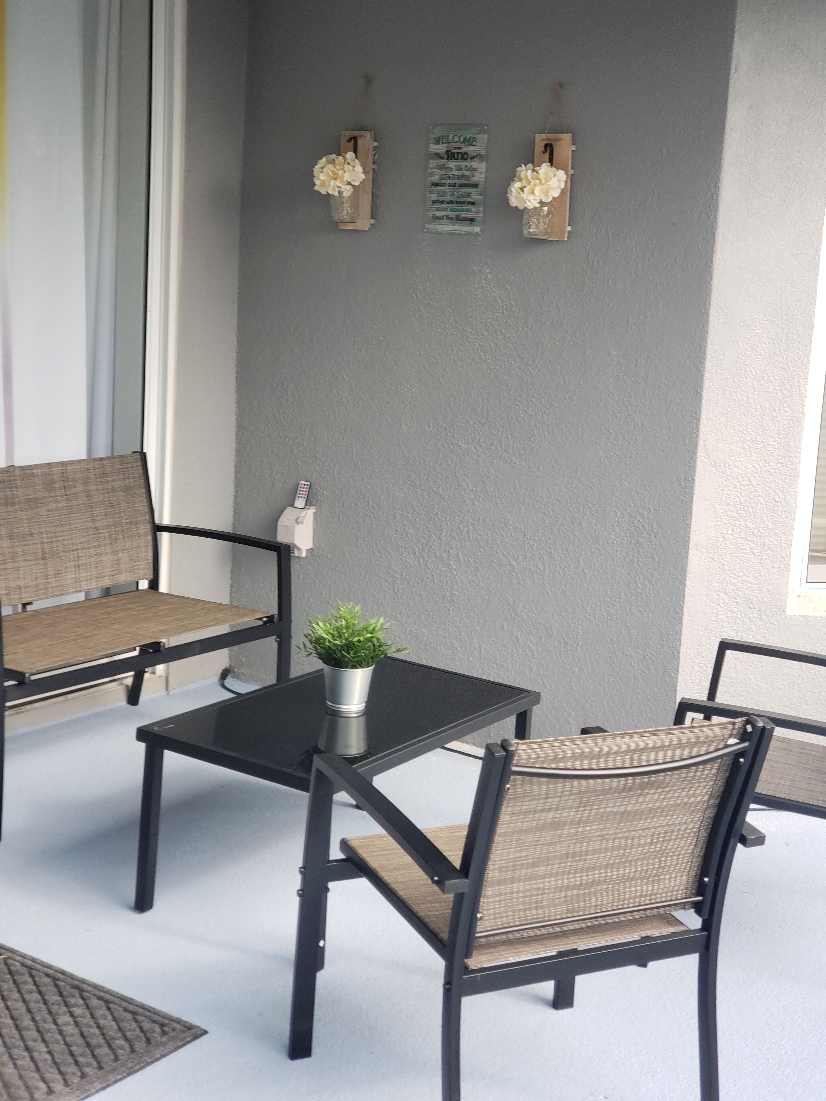 A patio area with two wicker chairs, a small table, and a potted plant. There are two signs on the wall, one welcoming guests to the patio and the other listing amenities.