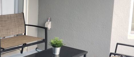 A patio area with two wicker chairs, a small table, and a potted plant. There are two signs on the wall, one welcoming guests to the patio and the other listing amenities.