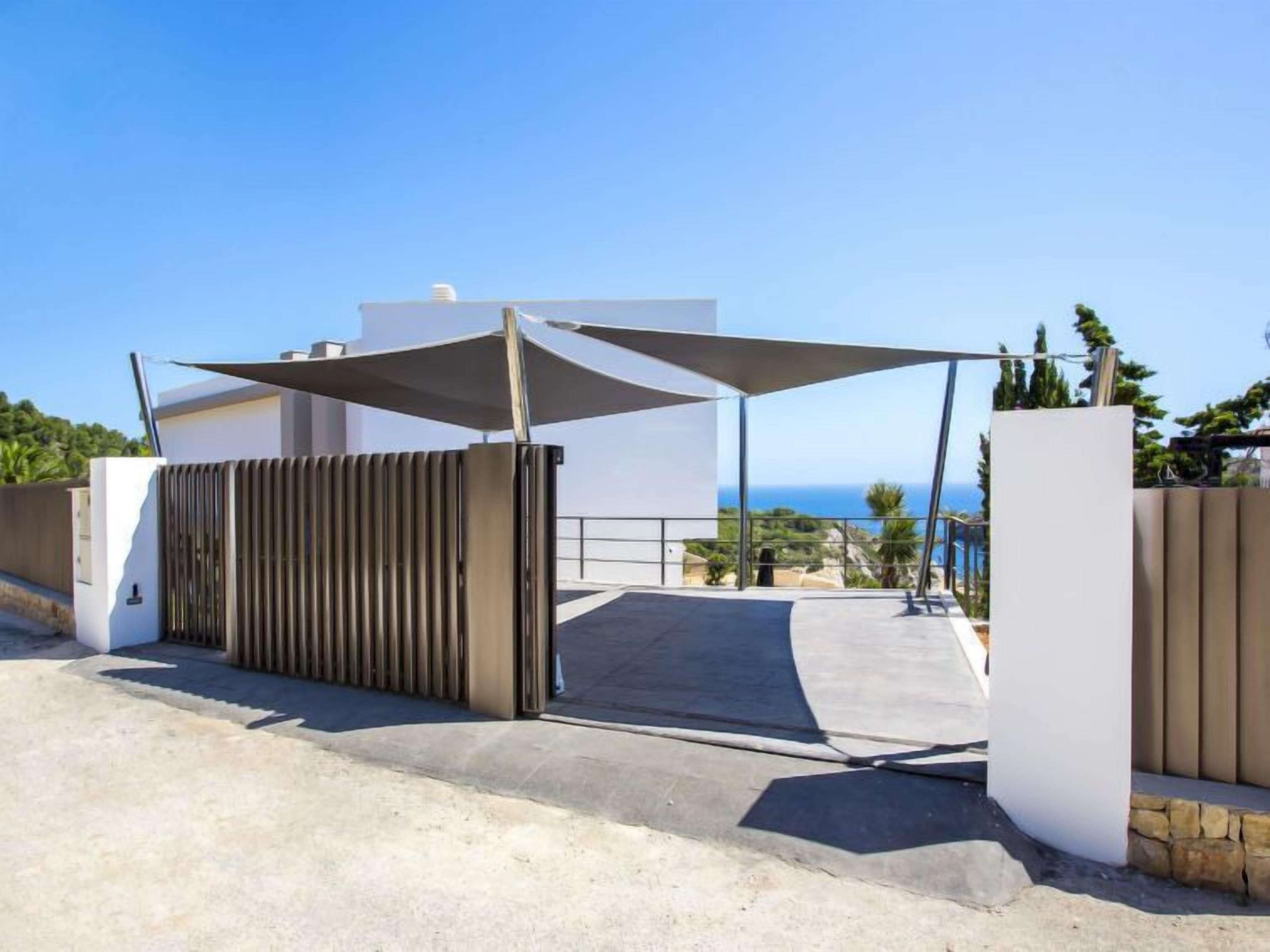 A gated entrance with a modern design, a clear view of the sea, and a building with a white facade in the background.