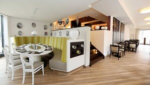 Een moderne eethoek met een ronde tafel, witte stoelen en een gele tafelkleed. Een trap leidt naar een balkon met een leuning.