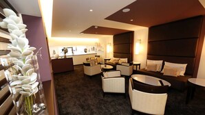 Een moderne hotelkamer met een donker hoofdbord, een zithoek met sofa’s en fauteuils, een klein rond tafeltje en een decoratieve vaas met witte bloemen.