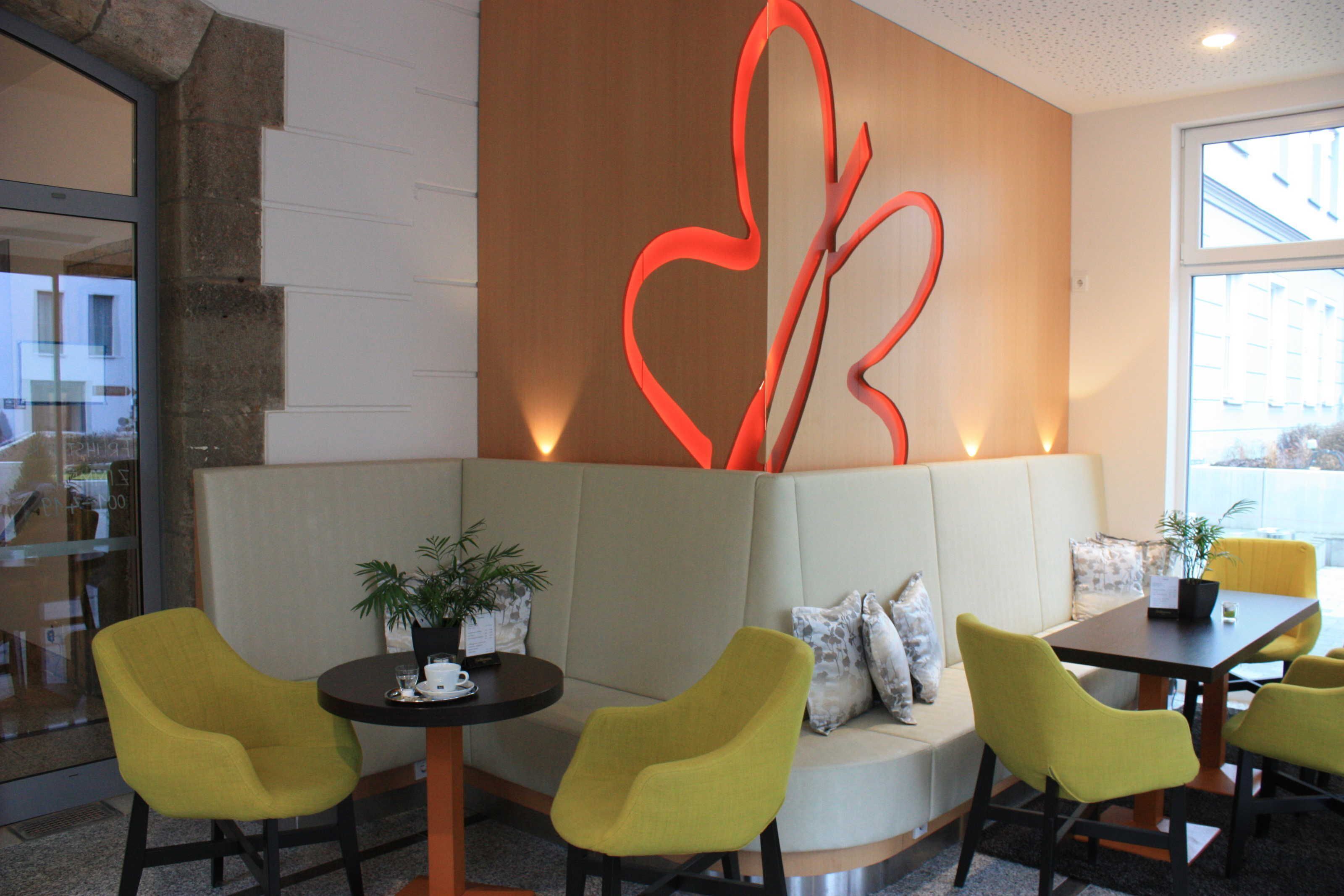 Ein modernes Interieur mit einer Sitzecke, die gelbe Stühle und ein Sofa umfasst, eine Holzvertäfelung mit rotem abstrakten Muster sowie ein kleiner Tisch mit einer Pflanze.