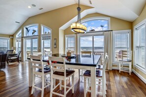 A dining area with a black table and white chairs, large windows with blinds, and a view of the ocean.