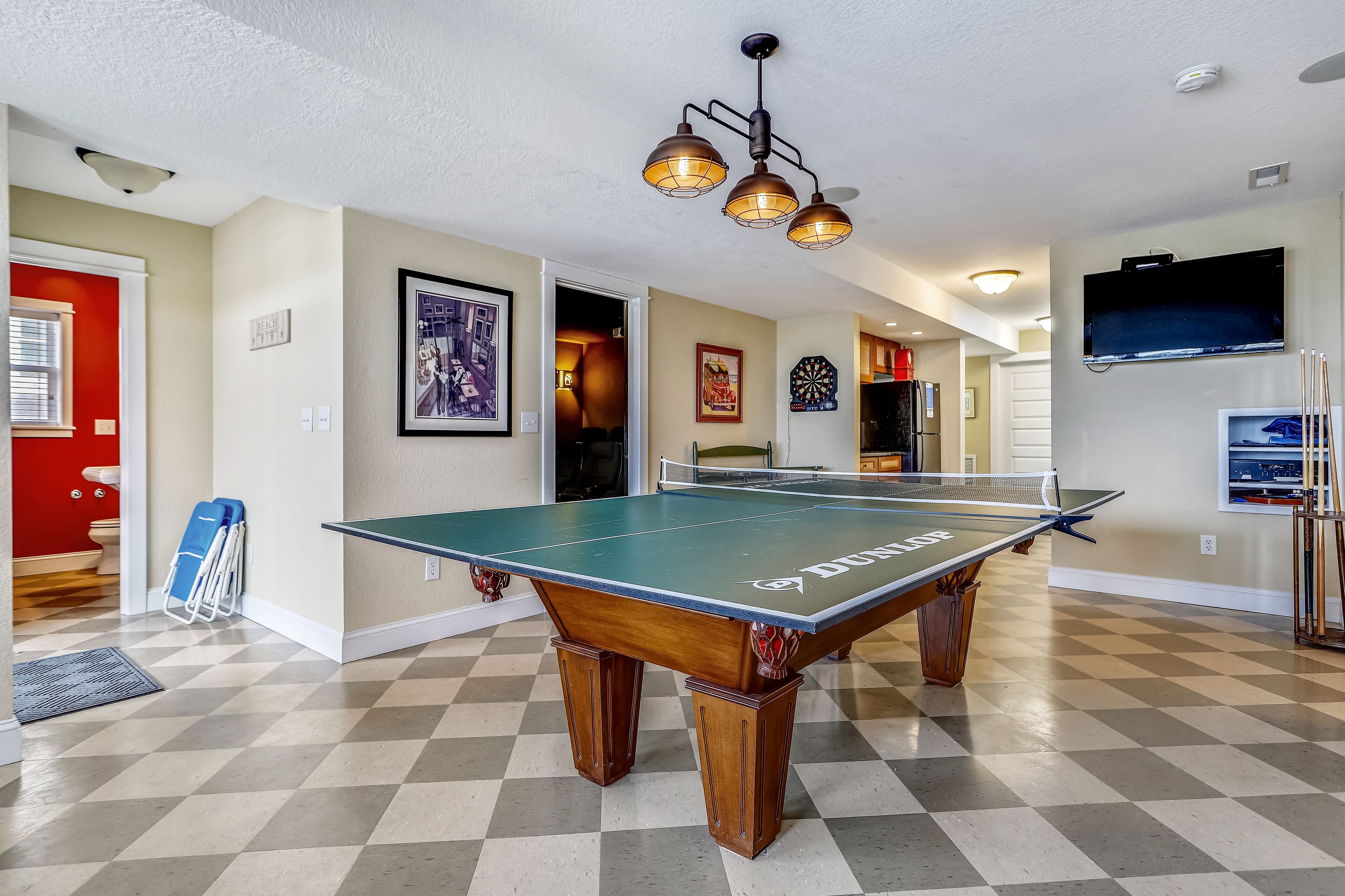 A ping pong table in a room with a checkered floor, a ceiling light fixture, and a television mounted on the wall.