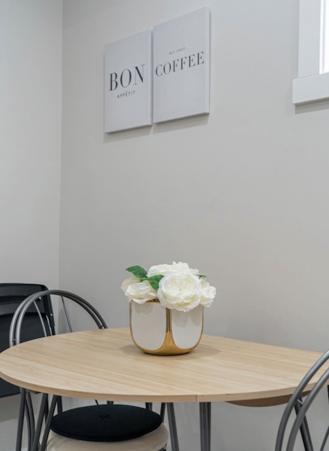 A round table with a white vase containing white flowers, two framed signs on the wall, and a black chair.