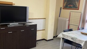 A room with a dark wooden cabinet, a flat-screen TV on top, a table with a patterned tablecloth, and a radiator in the background.