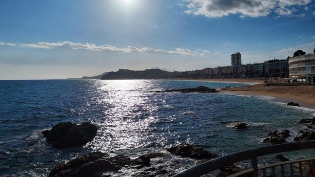 A coastal scene with a clear sky, a beach, and buildings in the background.