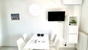 Ein moderner, minimalistischer Raum mit einem weißen Tisch, vier Stühlen, einem Flachbildfernseher, einer runden Leuchte und einer weißen Regaleinrichtung mit einem Bonsai-Baum.