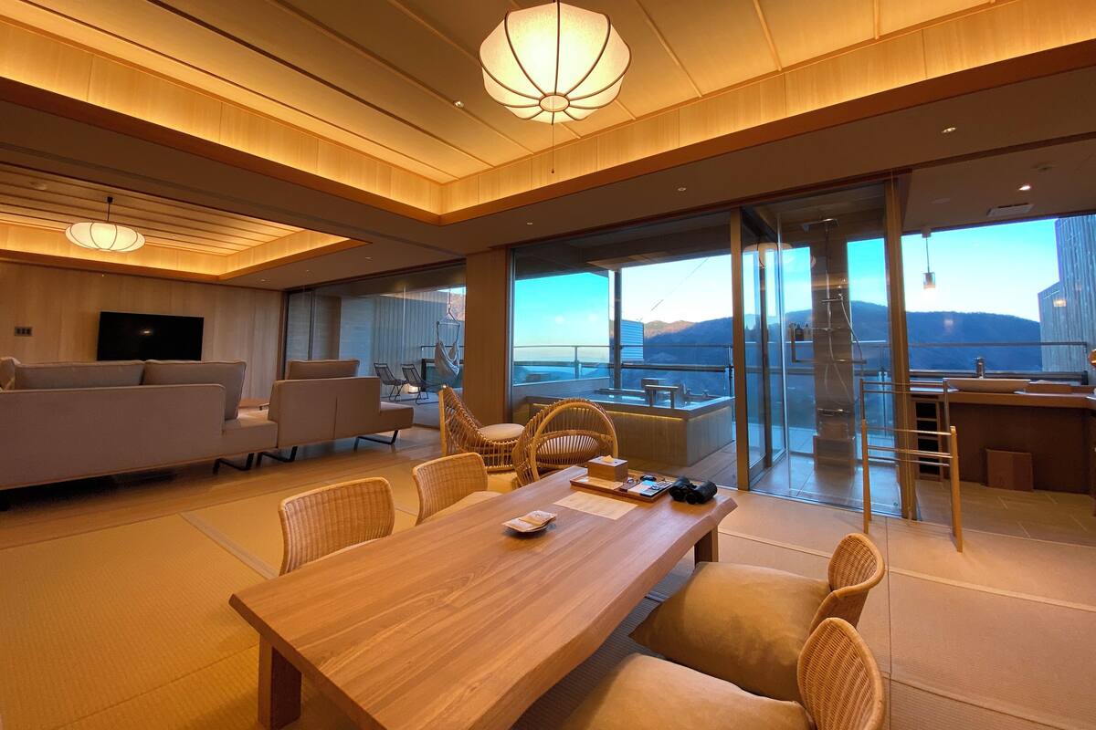 A modern living room with a wooden dining table, comfortable seating, a flat-screen TV, and a large window offering a view of the sea.