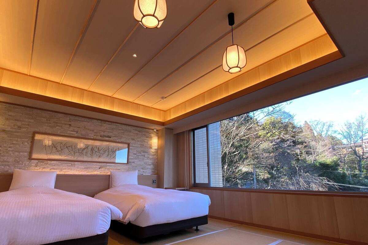 A hotel room with two beds, a large window with a view of trees, and a wooden ceiling.
