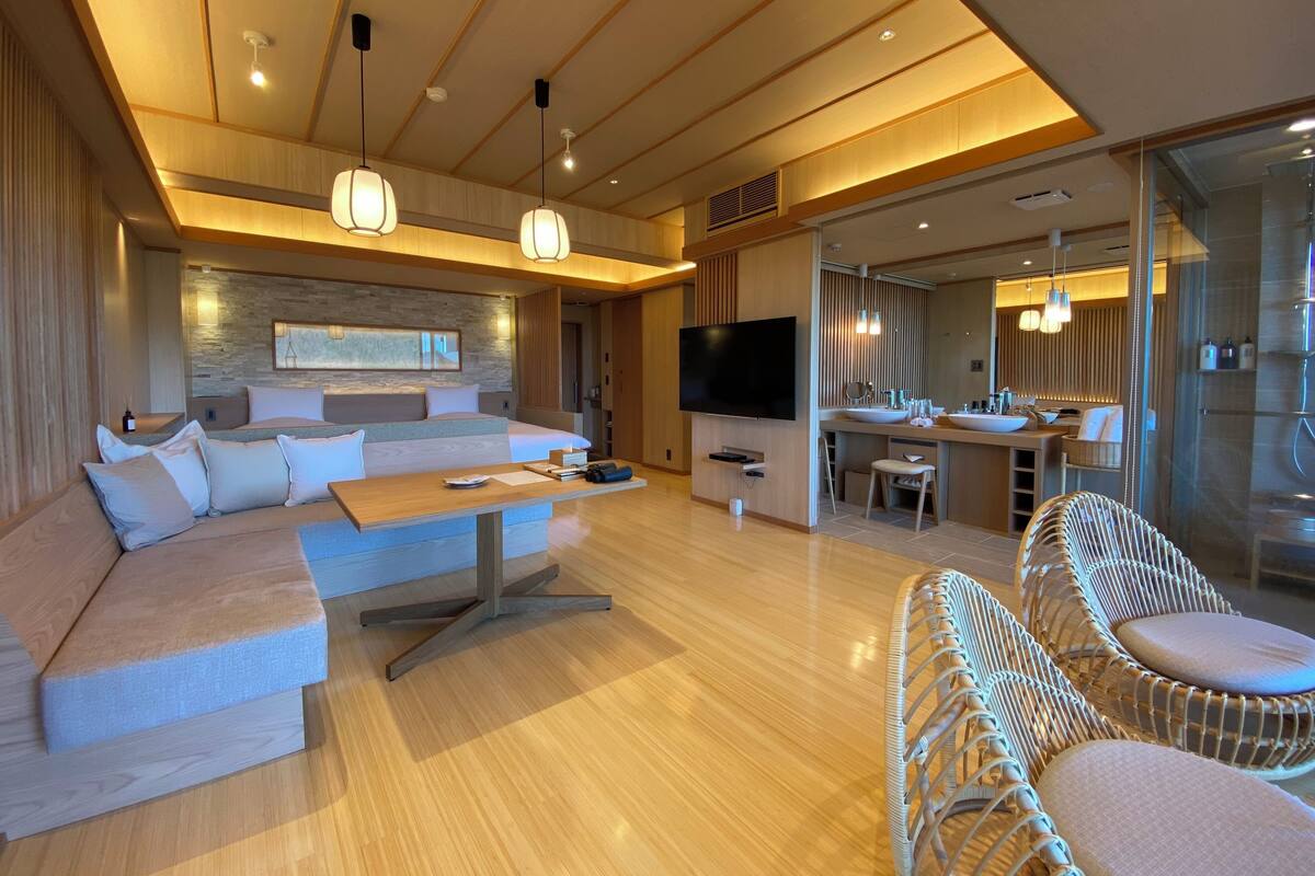 A modern hotel room with a large sofa, a dining area, and a TV.