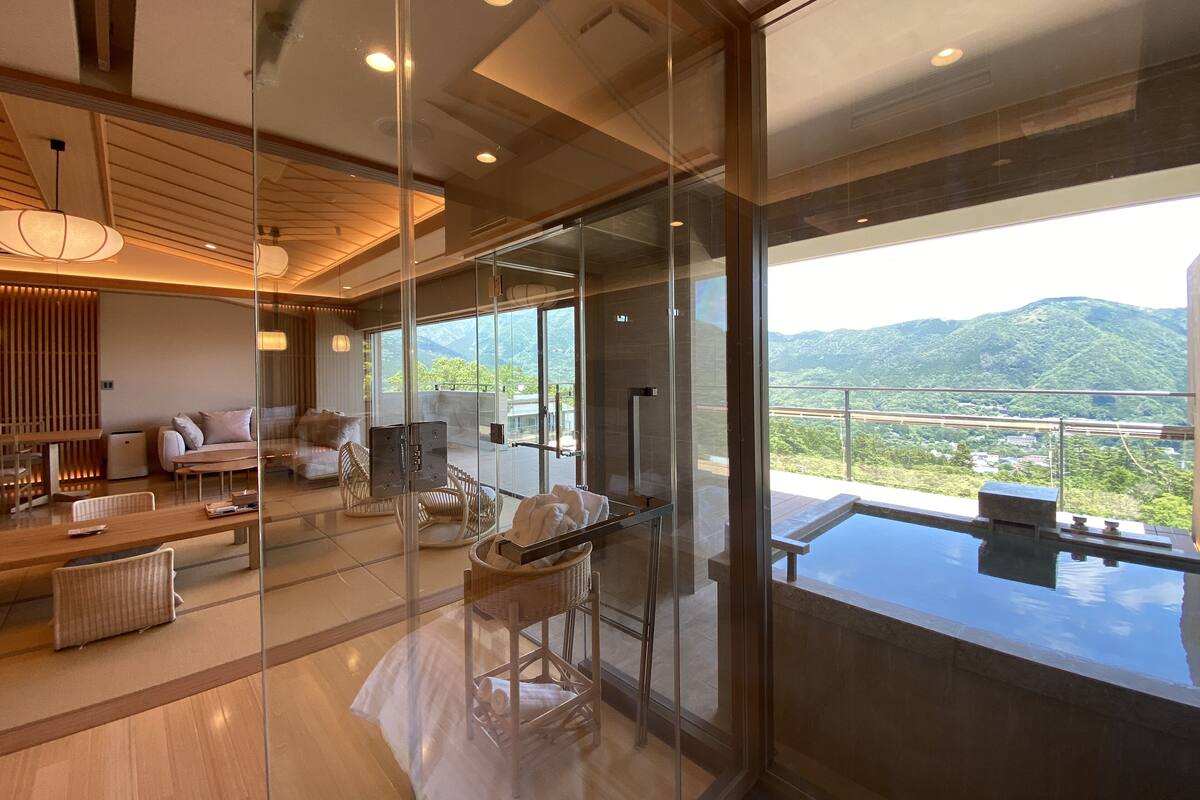 A modern hotel room with a glass wall, a balcony with a pool, and a view of mountains.