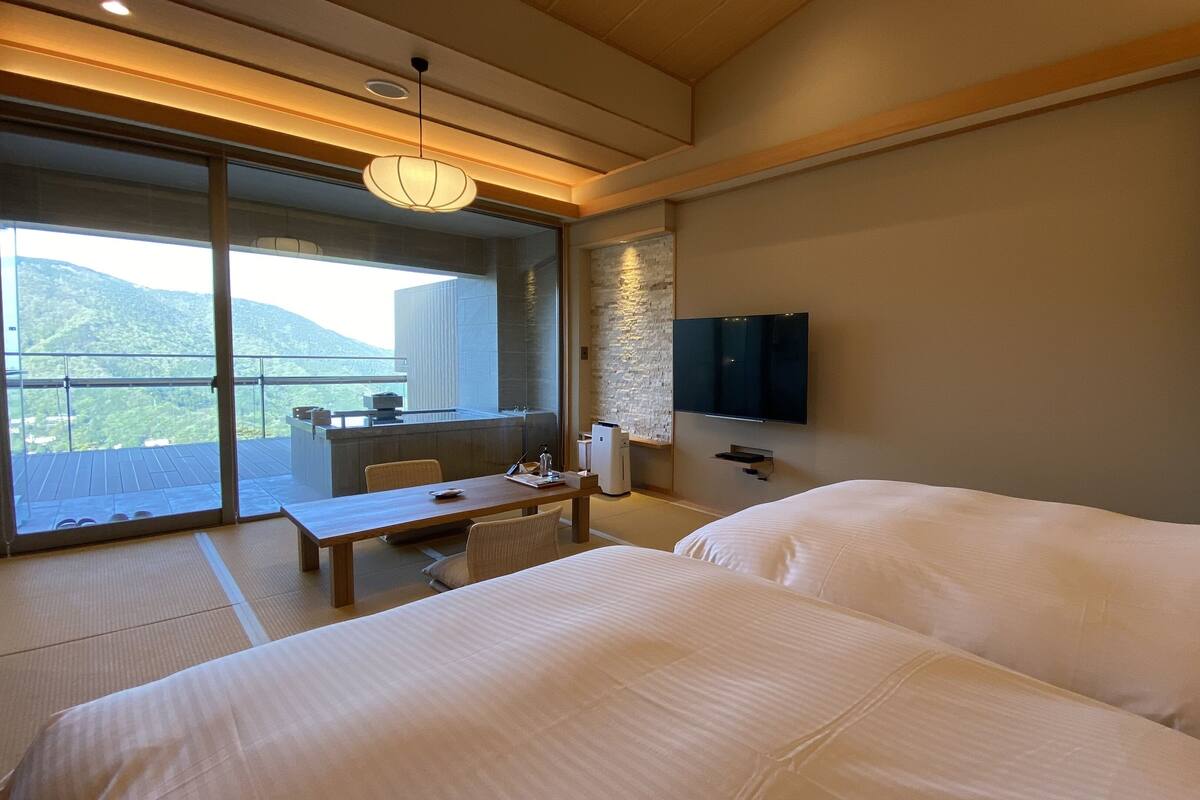 A hotel room with a large bed, a desk with a chair, a television mounted on the wall, and a balcony with a view of the mountains.