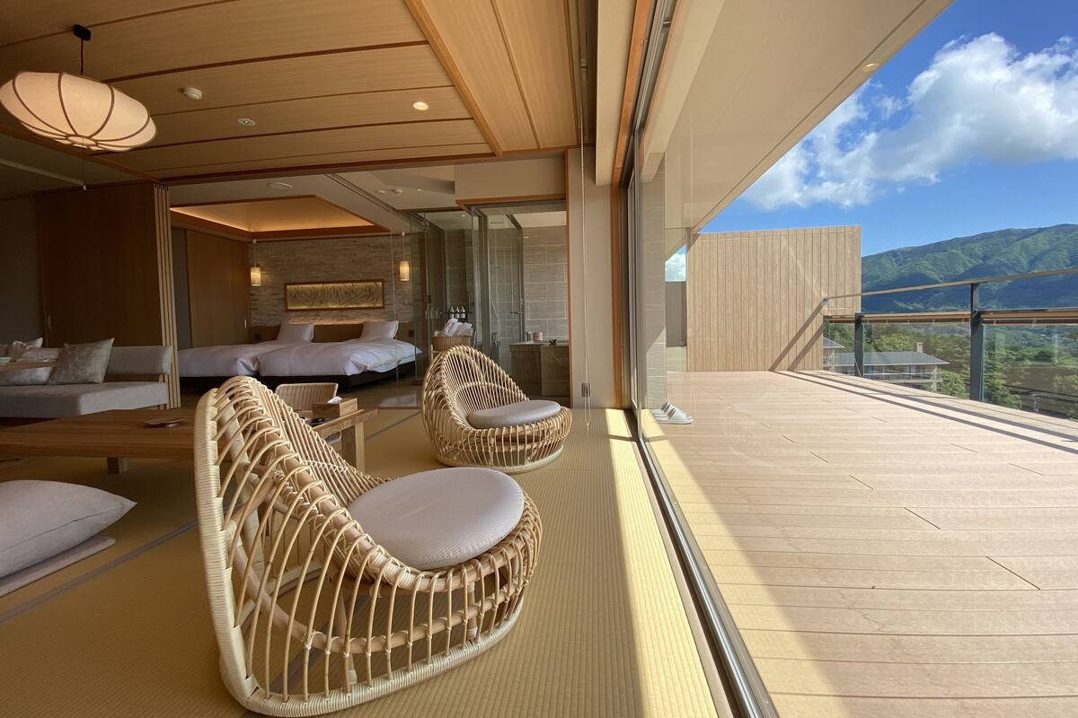 A modern hotel room with a large bed, two wicker chairs, a sofa, and a view of the mountains.