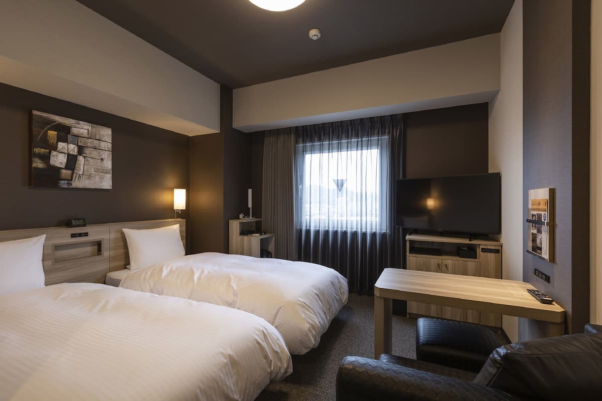 A hotel room with two beds, a desk, a television, and a window with curtains.