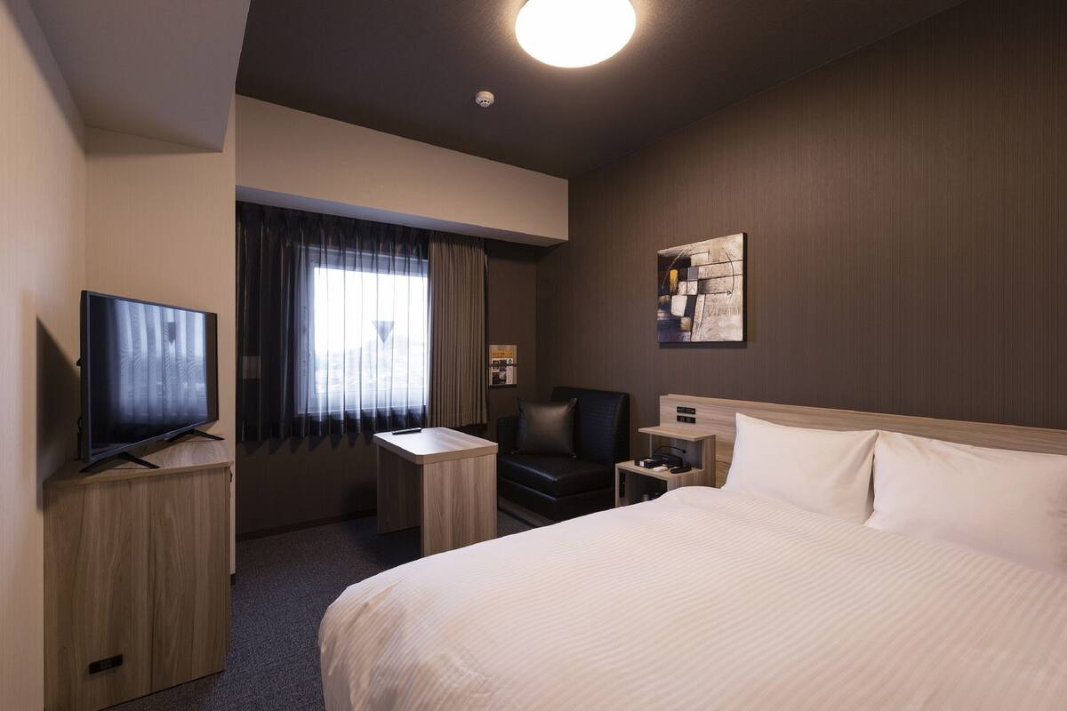 A hotel room with a large bed, a desk with a TV, a chair, a nightstand, and a window with curtains.