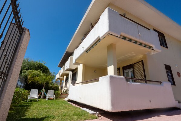 A two-story building with a flat roof, a balcony on the upper floor, and a small garden area with two white chairs.