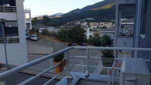 A balcony with a view of a parking area, a building with a white facade, and a mountainous landscape in the background.