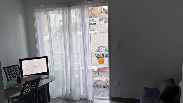 A living room with a sofa, a dining table with chairs, and a laptop. There is a sliding glass door leading to an outdoor area with a car and a plant.