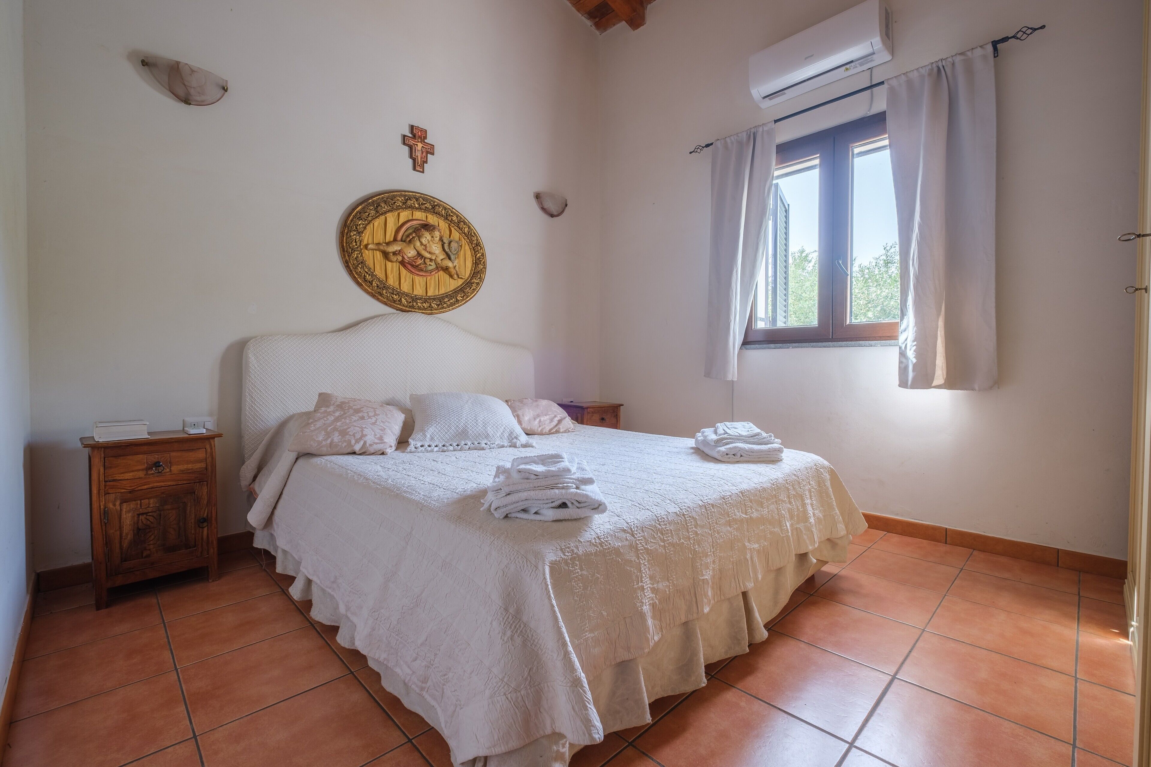 A bedroom with a bed, a nightstand, a wall decoration, a window with curtains, and a tiled floor.