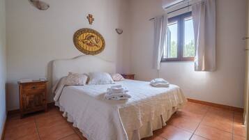 A bedroom with a bed, a nightstand, a wall decoration, a window with curtains, and a tiled floor.