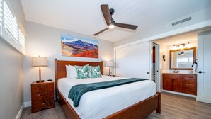 A bedroom with a wooden bed, a ceiling fan, a nightstand, a lamp, a painting, and a bathroom with a mirror and cabinet. - Big Ocean View from your Sugar Beach Retreat -King Bed, Central AC, WiFi  #208 (Kihei)