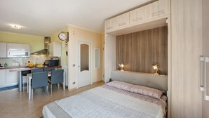 Ein Schlafzimmer mit Bett, Schreibtisch mit Stühlen, einer Küchenzeile und einer Uhr an der Wand.