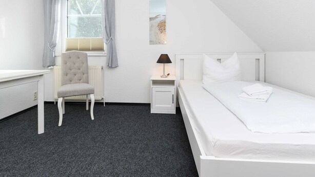 Een slaapkamer met een bed, een stoel, een nachtkastje, een lamp en een raam met gordijnen.